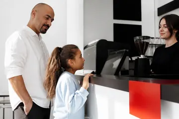 Dokuz yaşında bir kız çocuğu bir kafenin tezgâhında cesaret adımı olarak kendi siparişini verirken babası arkasında destek olarak duruyor